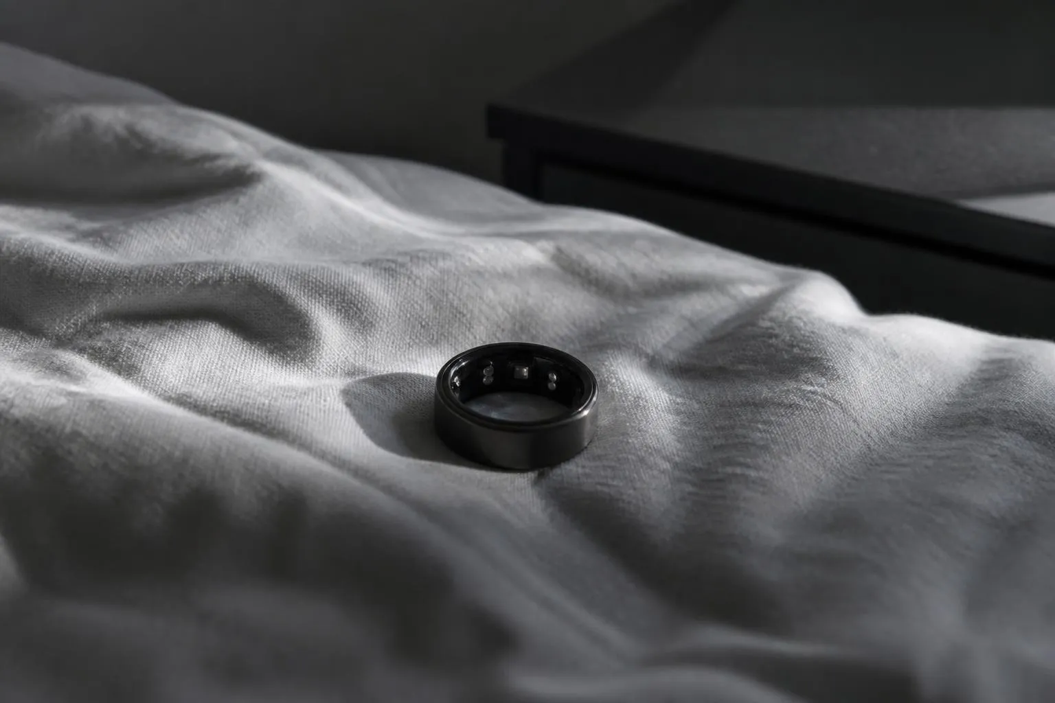 Bague connectée retournée sur un drap froissé, capteurs vers le haut, dans la lumière grise du petit matin