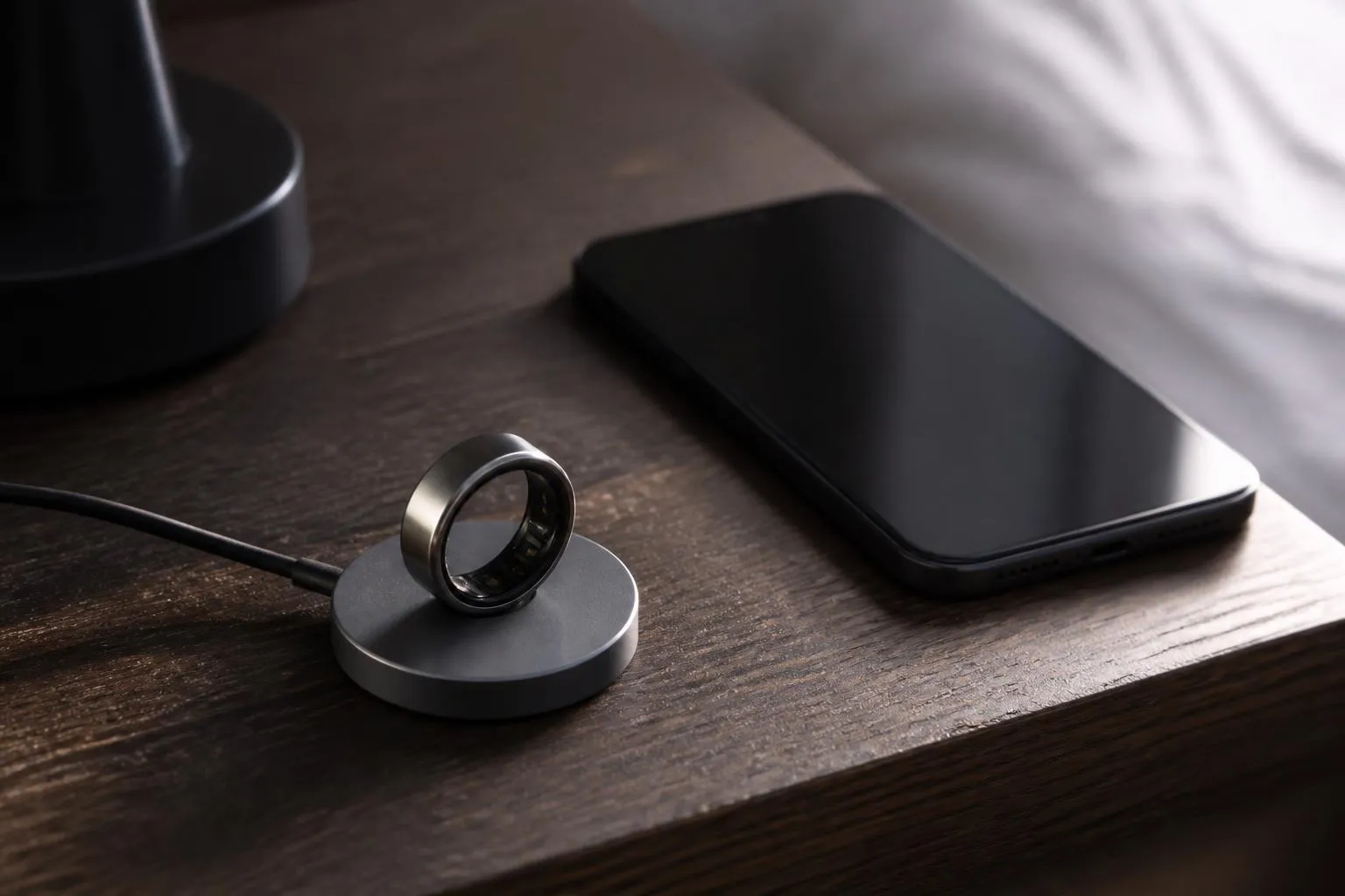 Bague connectée posée sur son socle de charge à côté d'un téléphone éteint, sur une table sombre
