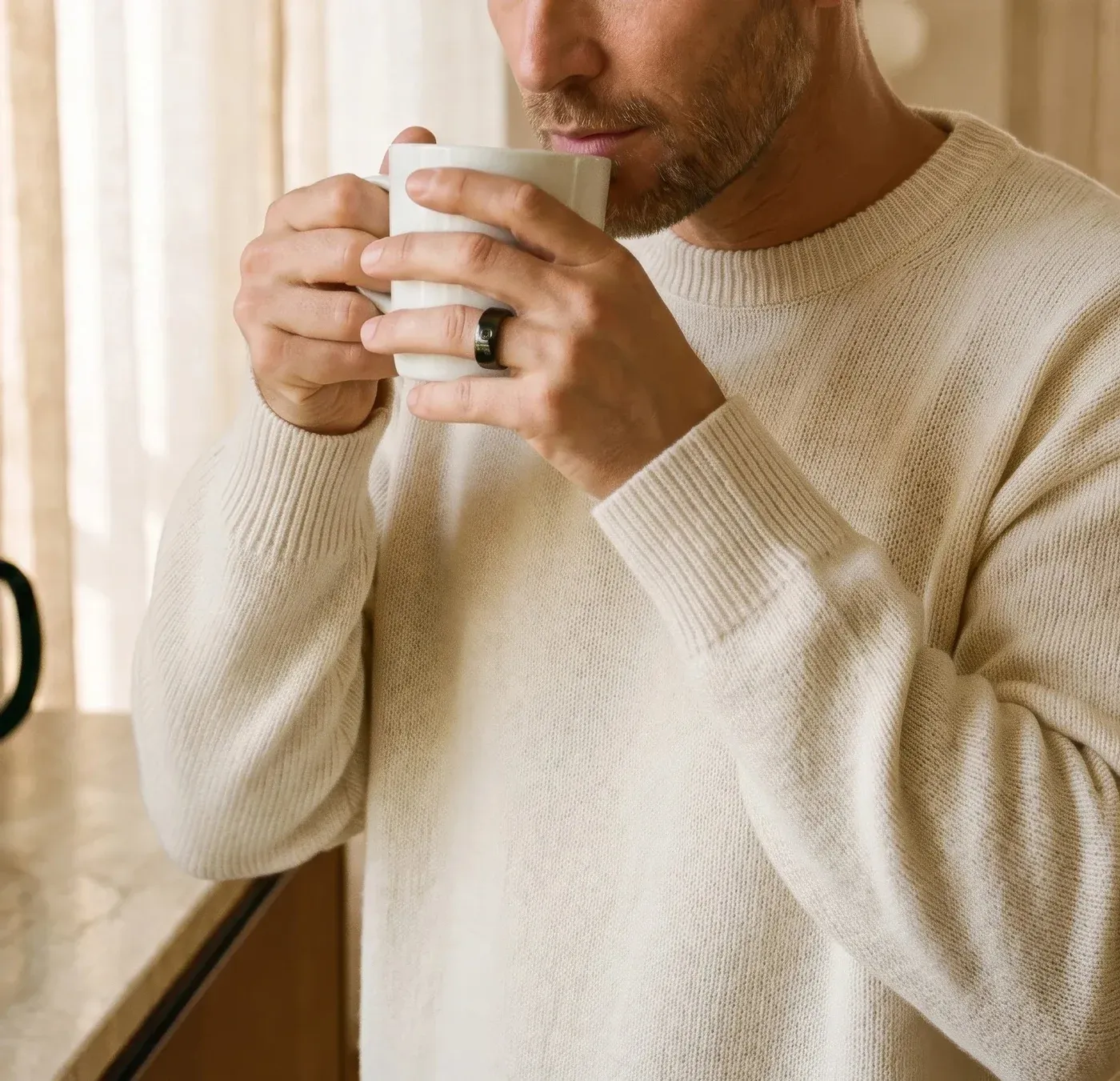 Un homme en pull écru tient une tasse à deux mains, la Circular Ring 2 noire à l'annulaire